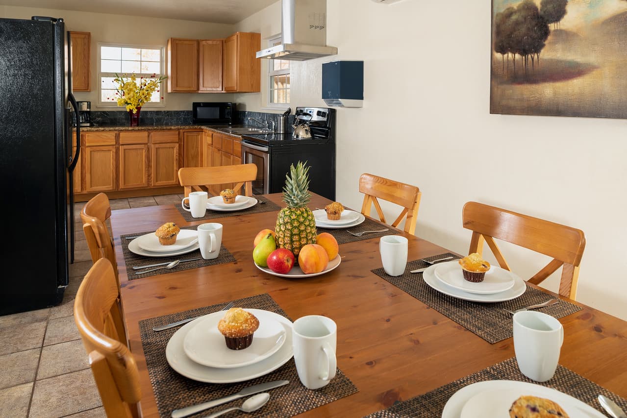 Large dining room table with chairs set for breakfast with full kitchen with refrigerator and stove in background