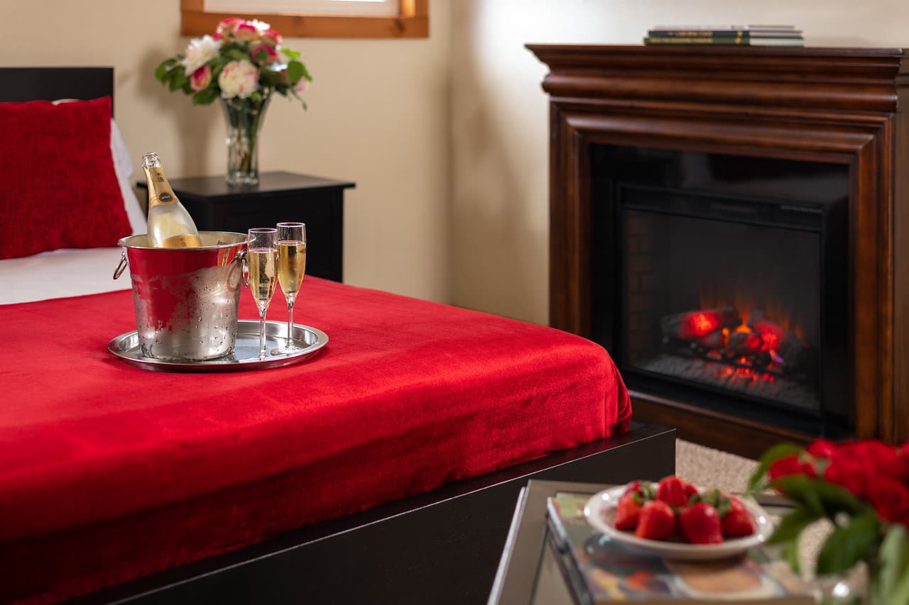 Fireplace, silver platter with ice bucket, champagne, and two glasses on bed with fresh strawberries and flowers on table nearby