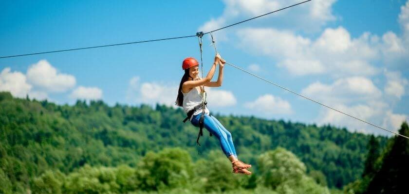 A woman in a red helmet zip lines through a green landscape under a blue sky.