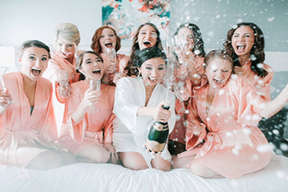 8 happy bridesmaids in peach robes kneeling on a bed with champagne glasses in hand, and bride in the middle in a white robe holding a champagne bottle and glass with confetti in the air.