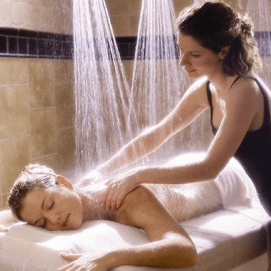 Woman laying on stomach on massage table under rain shower heads with a masseuse massaging her back