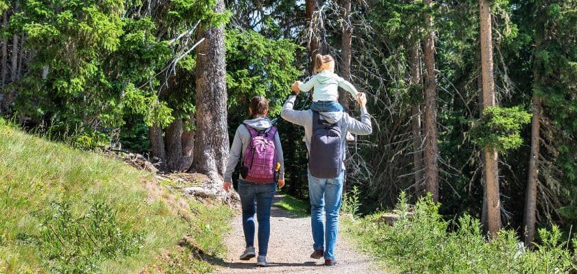 Two adults walk along a forest path, one carrying a child on their shoulders.