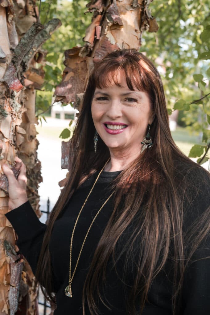 Anna Russell with long dark hair, bangs, and in a black top with greenery in the background