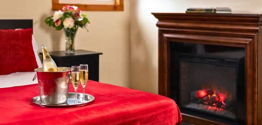 A cozy bedroom featuring a red bedspread, a fireplace, a bottle of champagne in an ice bucket, and two glasses.