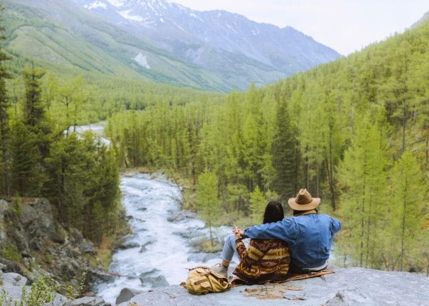 A couple sits on a rock by a river, surrounded by lush green mountains and forests.