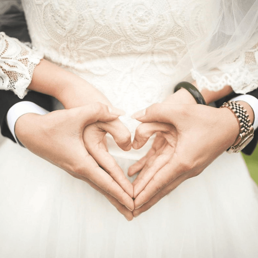 Groom with his arms around the bride dressed in wedding clothes, holding hands in the shape of a heart
